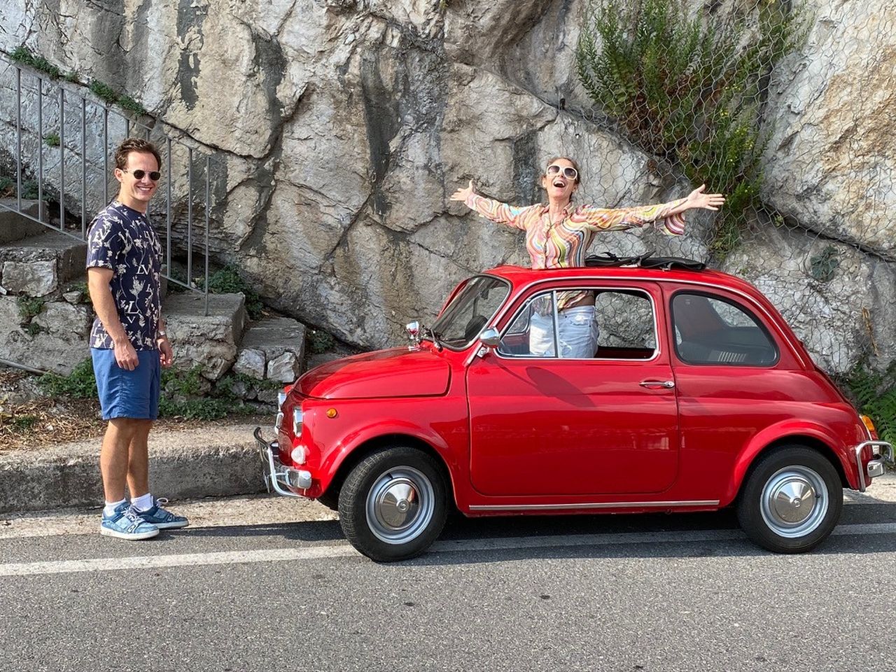 Red vintage car parked beside rocky wall and stairs.