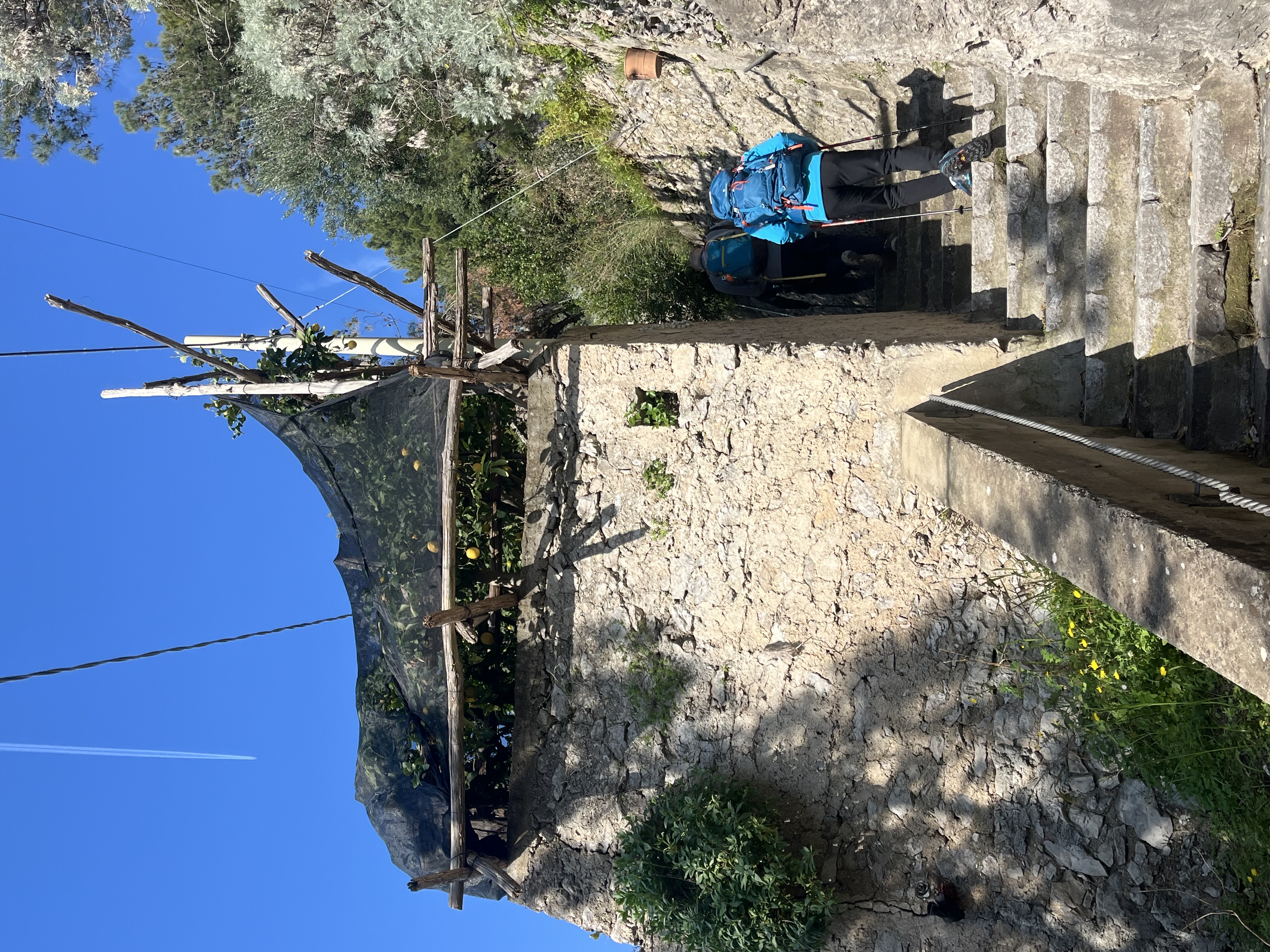 People climbing stone steps beside a rustic wall under a clear sky.