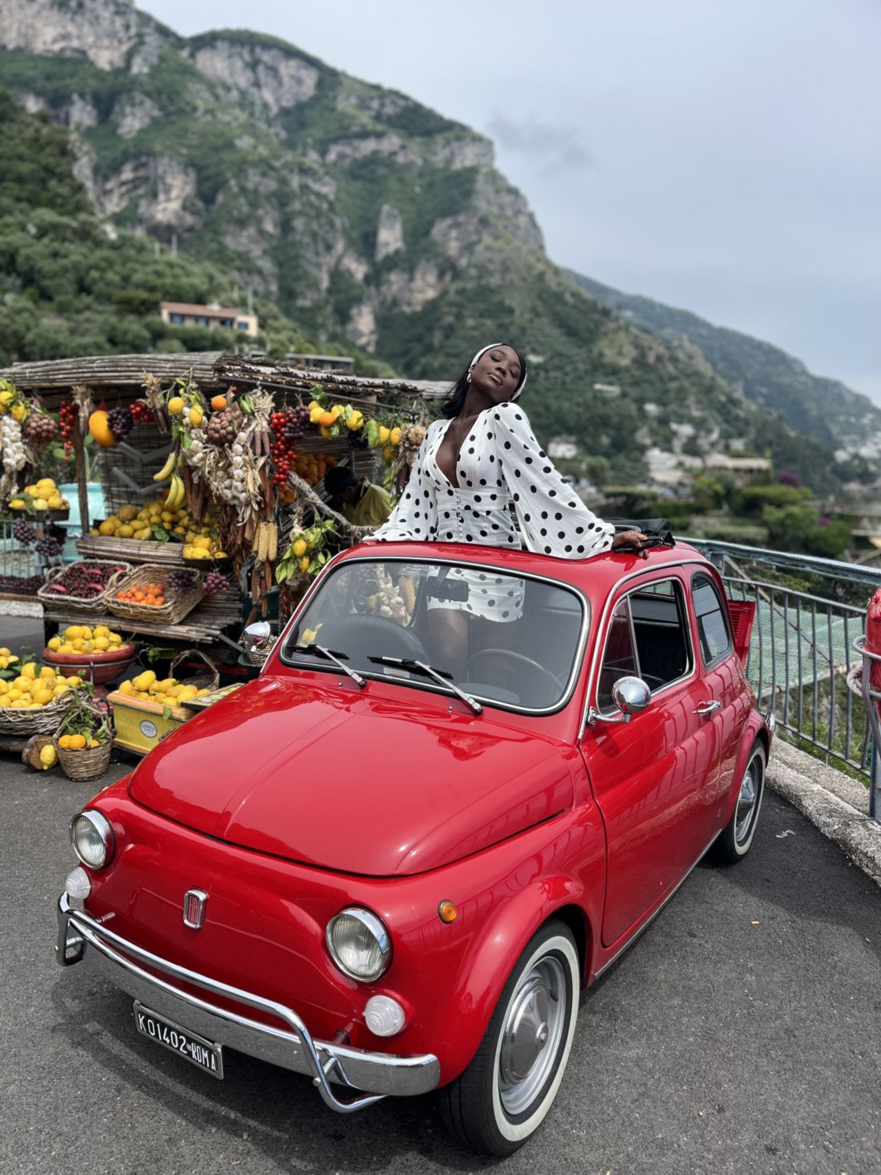 Red vintage car parked near fruit stand with scenic mountain backdrop.
