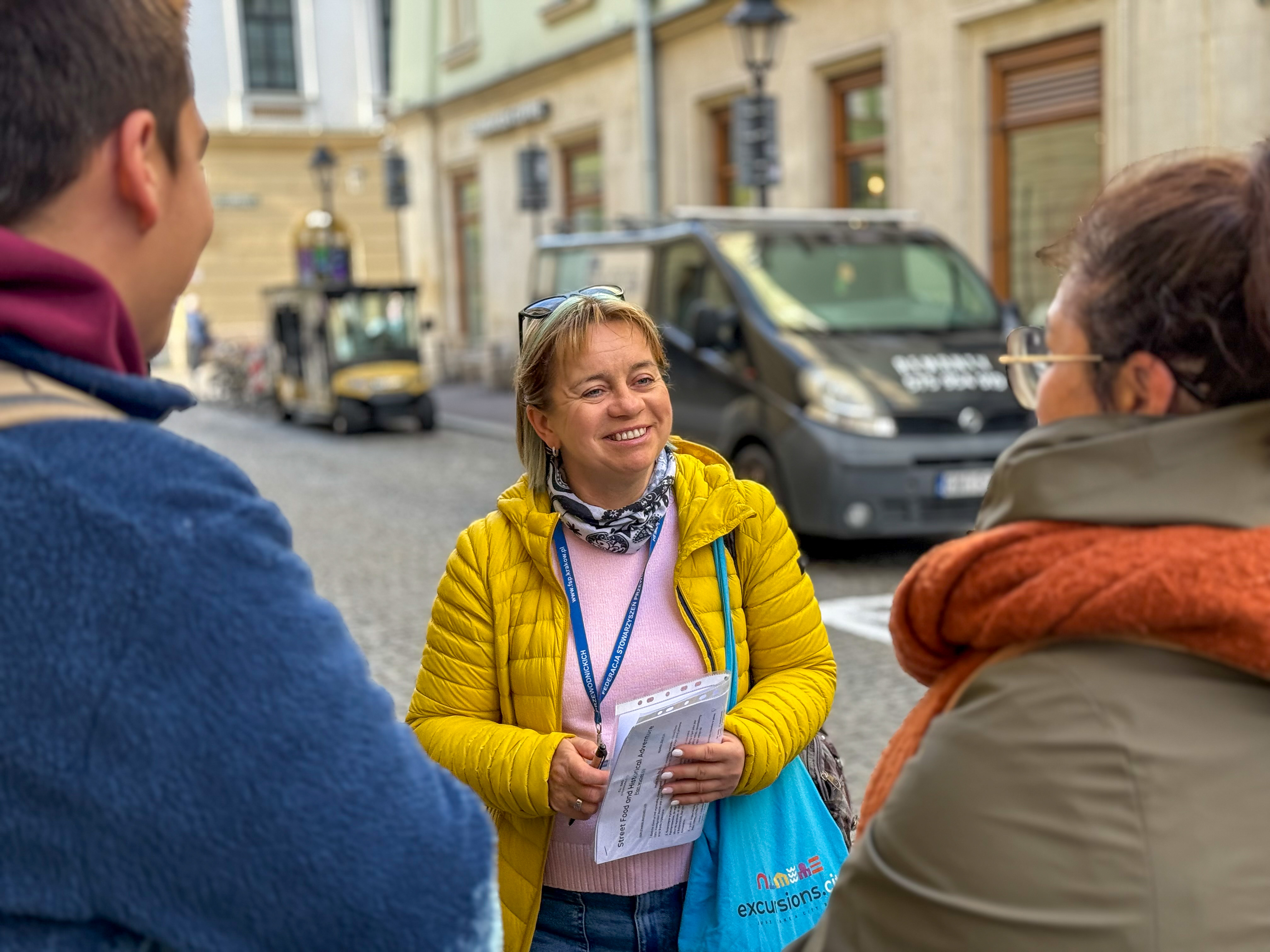 Person in yellow jacket talking to two people on a cobblestone street.