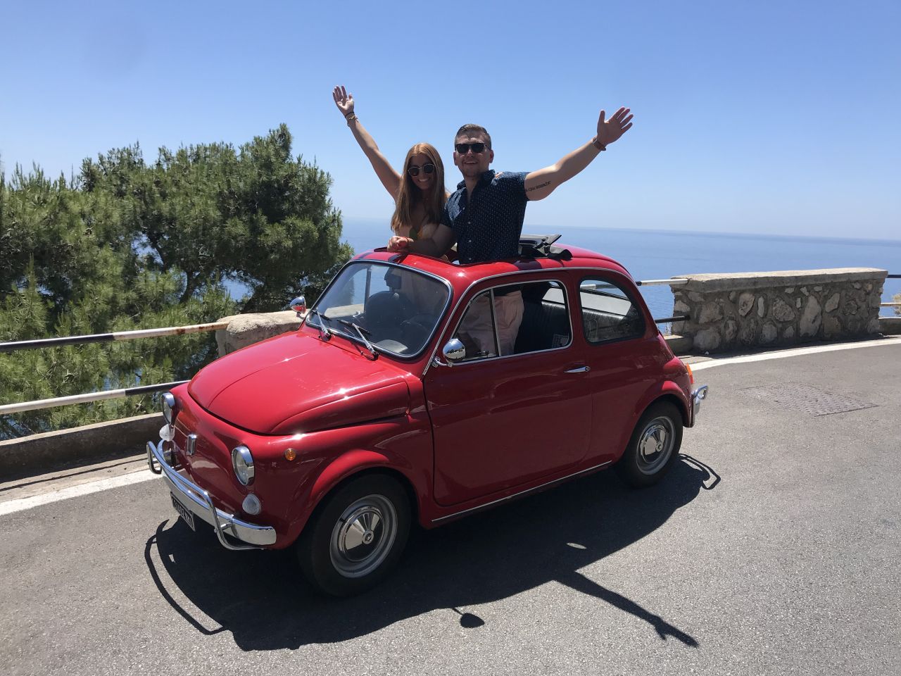 Two people in red vintage car with arms raised, ocean view.