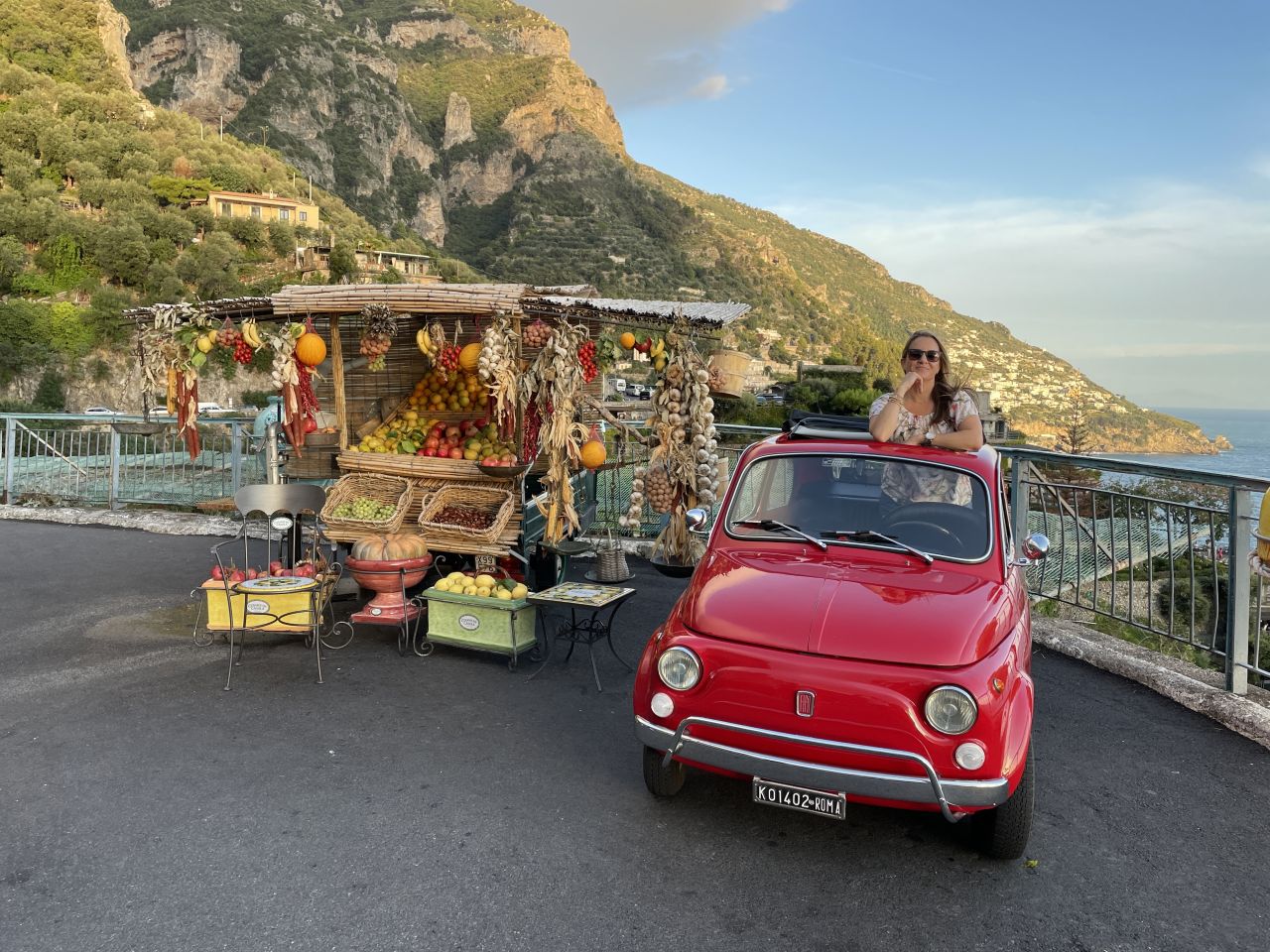 Red vintage car near fruit stand on coastal road.