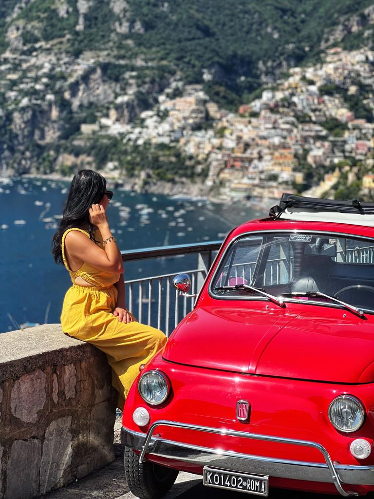 Woman in yellow dress sitting by red vintage car overlooking coastal town.