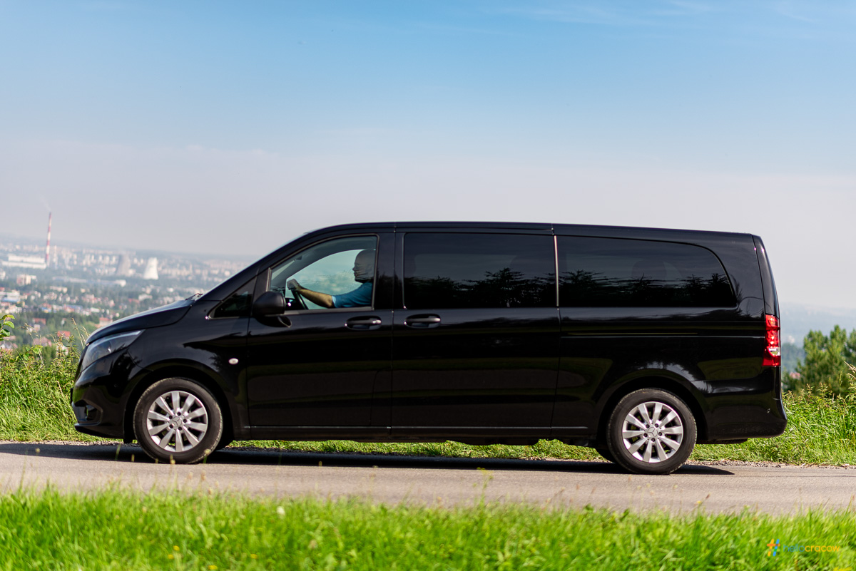 Black van driving on a scenic road with green grass and blue sky.