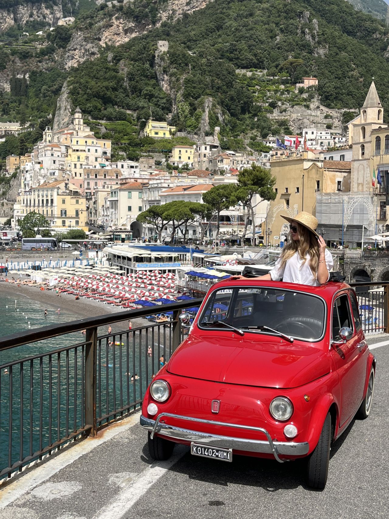 Red vintage car parked by seaside town with colorful buildings.