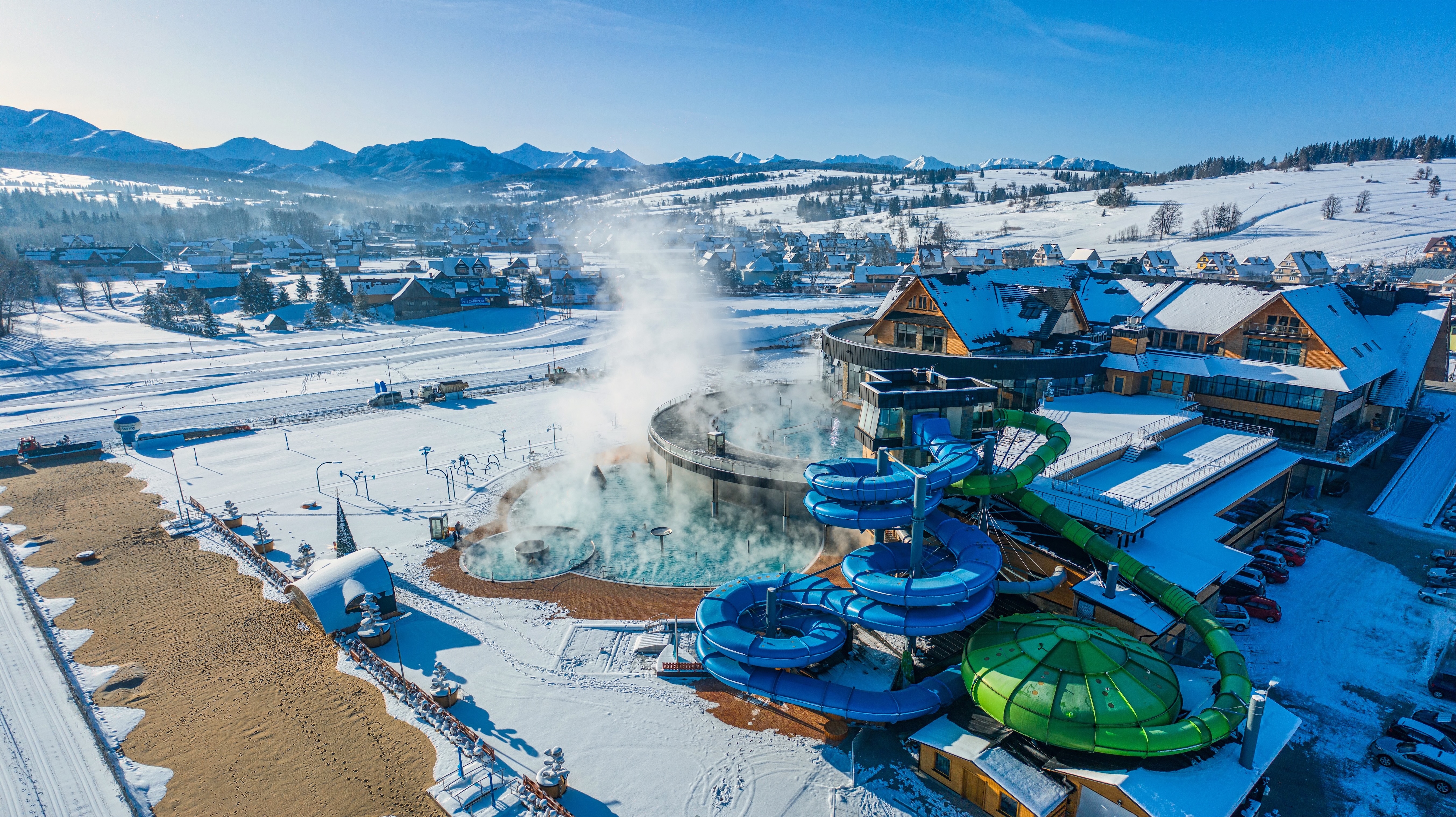 Aerial view of snowy water park with slides and steaming pools, surrounded by mountains.