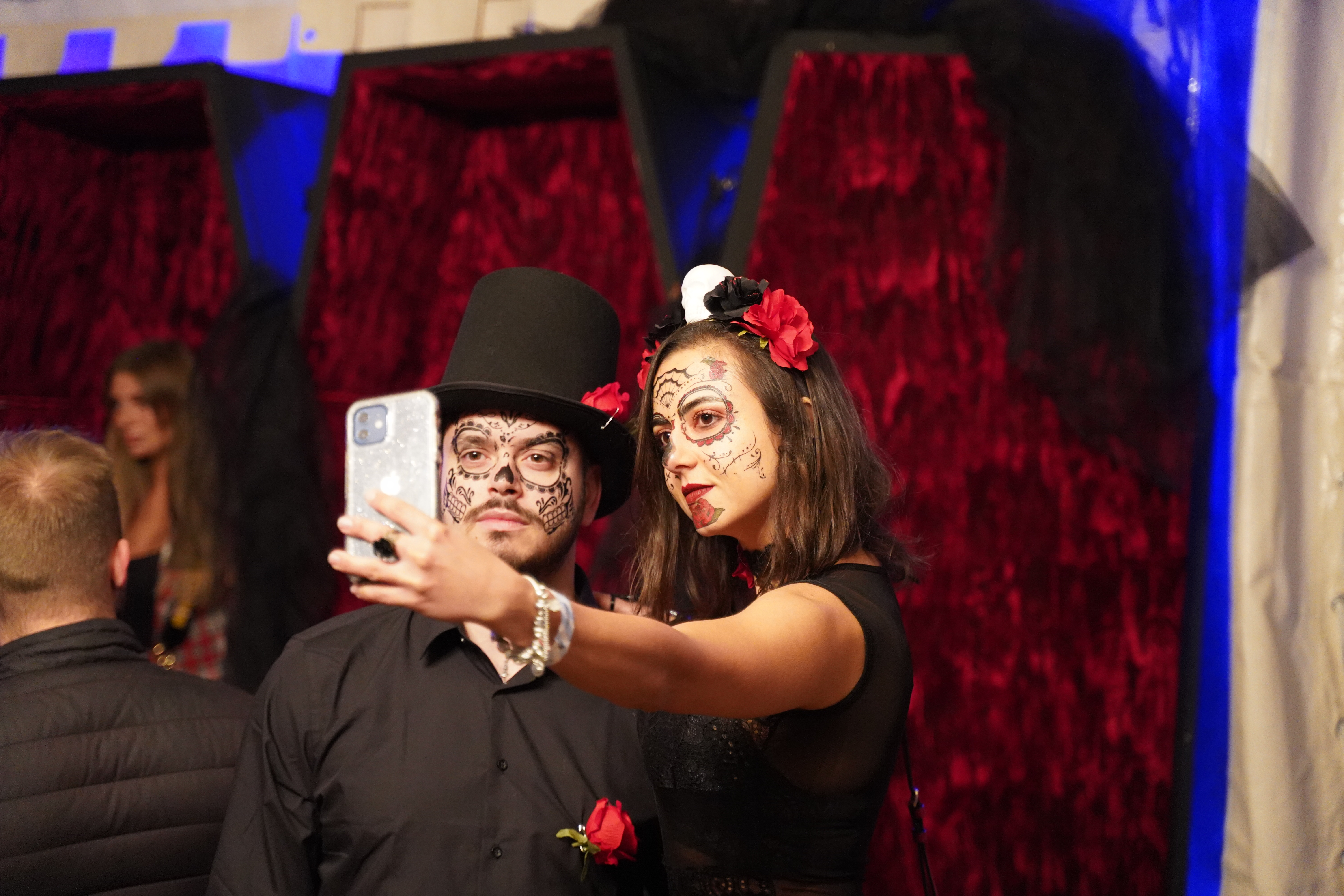 Couple wearing sugar‑skull facepaint and floral accessories taking a selfie at lively indoor celebration.